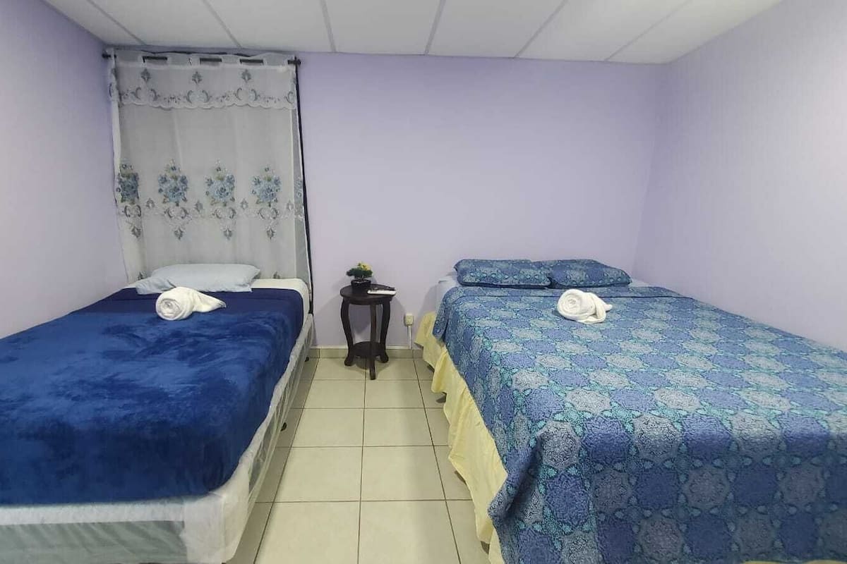 Een kamer met twee eenpersoonsbedden, elk met een blauwe spreidek en witte kussens. Daartussen staat een klein tafeltje met een plant.