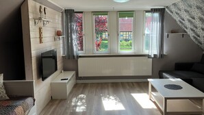 A modern living room with a sofa, a TV mounted on a wooden wall, a dining table, and a window with curtains. - Small, fine vacation apartment in the heart of Fürstenau (Fürstenau)
