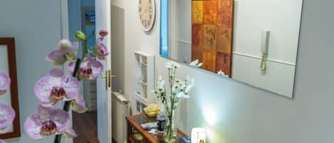 A hallway with a wooden door, a white radiator, a glass table with a vase of flowers, and a mirror reflecting a painting.
