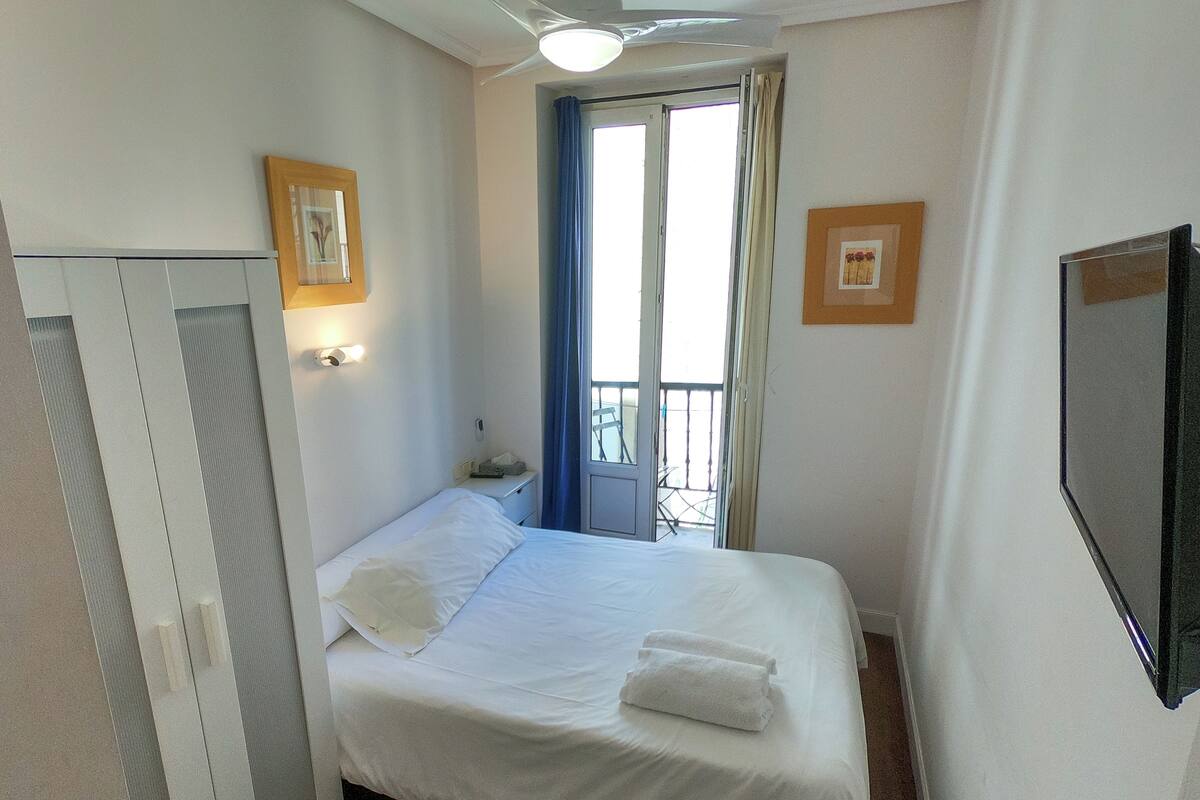 A single bed with white linens, a bedside table with a lamp, a window with blue curtains, and a wall-mounted TV.