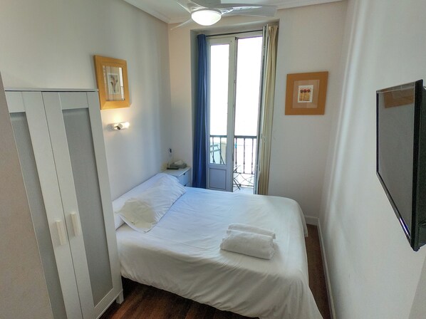 A single bed with white linens, a bedside table with a lamp, a window with blue curtains, and a wall-mounted TV. - Pension Anne (San Sebastián)