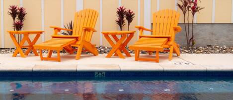 A poolside area with three orange Adirondack chairs and a sign reading 'Pelicans Roast #103 Bird in Paradise'.