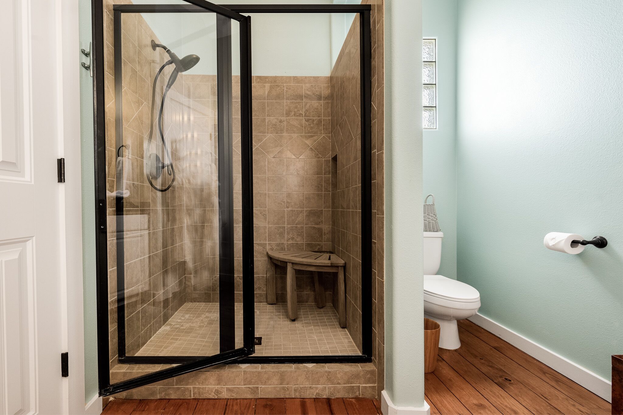 Een badkamer met een douche met glazen wanden, een houten krukje, een toilet en een witte deur.