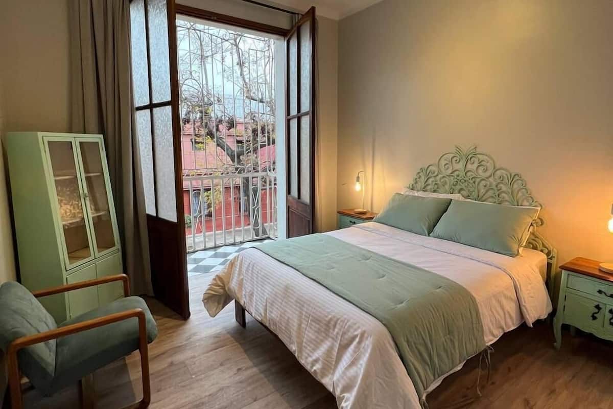 Une chambre à coucher avec un grand lit, une chaise, une armoire et une fenêtre donnant sur les bâtiments environnants.