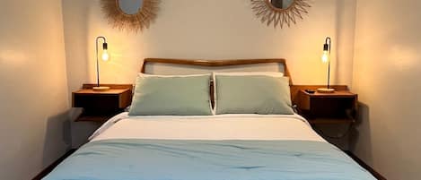 Una cama bien hecha con estructura de madera y ropa blanca, flanqueada por dos mesitas de noche con lámparas, y tres espejos colocados en la pared arriba.