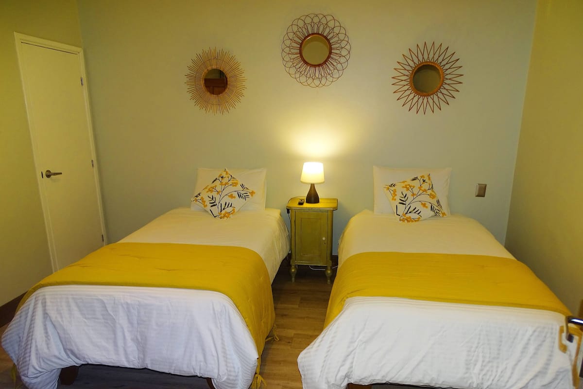 Une chambre avec deux lits, chacun recouvert d’une couvre-lit jaune et doté de draps blancs, une table de chevet avec une lampe, et des miroirs décoratifs en forme de soleil accrochés au mur.