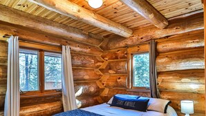A log cabin bedroom with a bed, two windows, and a nightstand. - Retreat at Blueberry (Osage)
