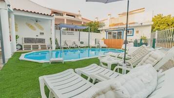 Una piscina con sillas de descanso y una sombrilla, rodeada de un césped cuidado y un edificio con tejado de baldosas.