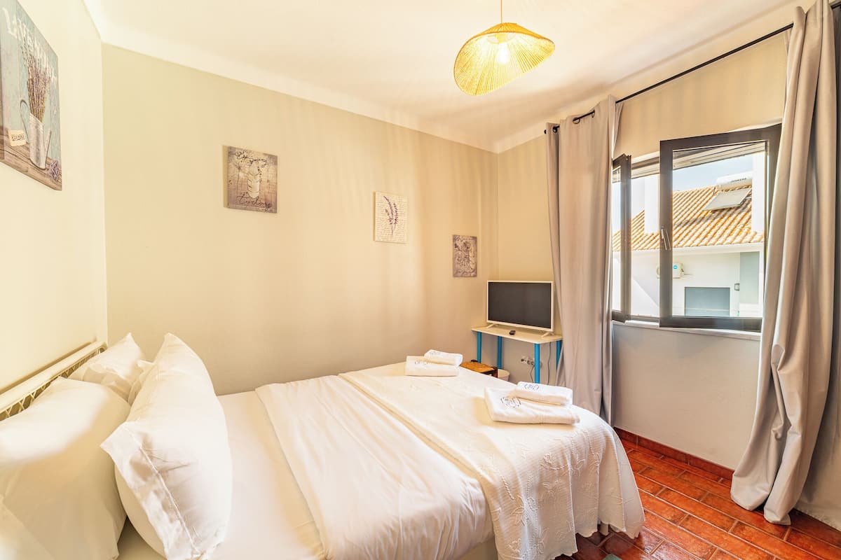 A hotel room with a bed, a television on a stand, a window with curtains, and artwork on the walls.