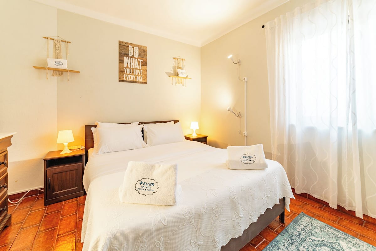 A hotel room with a large bed, two bedside tables with lamps, a wall-mounted shelf, and a window with curtains.