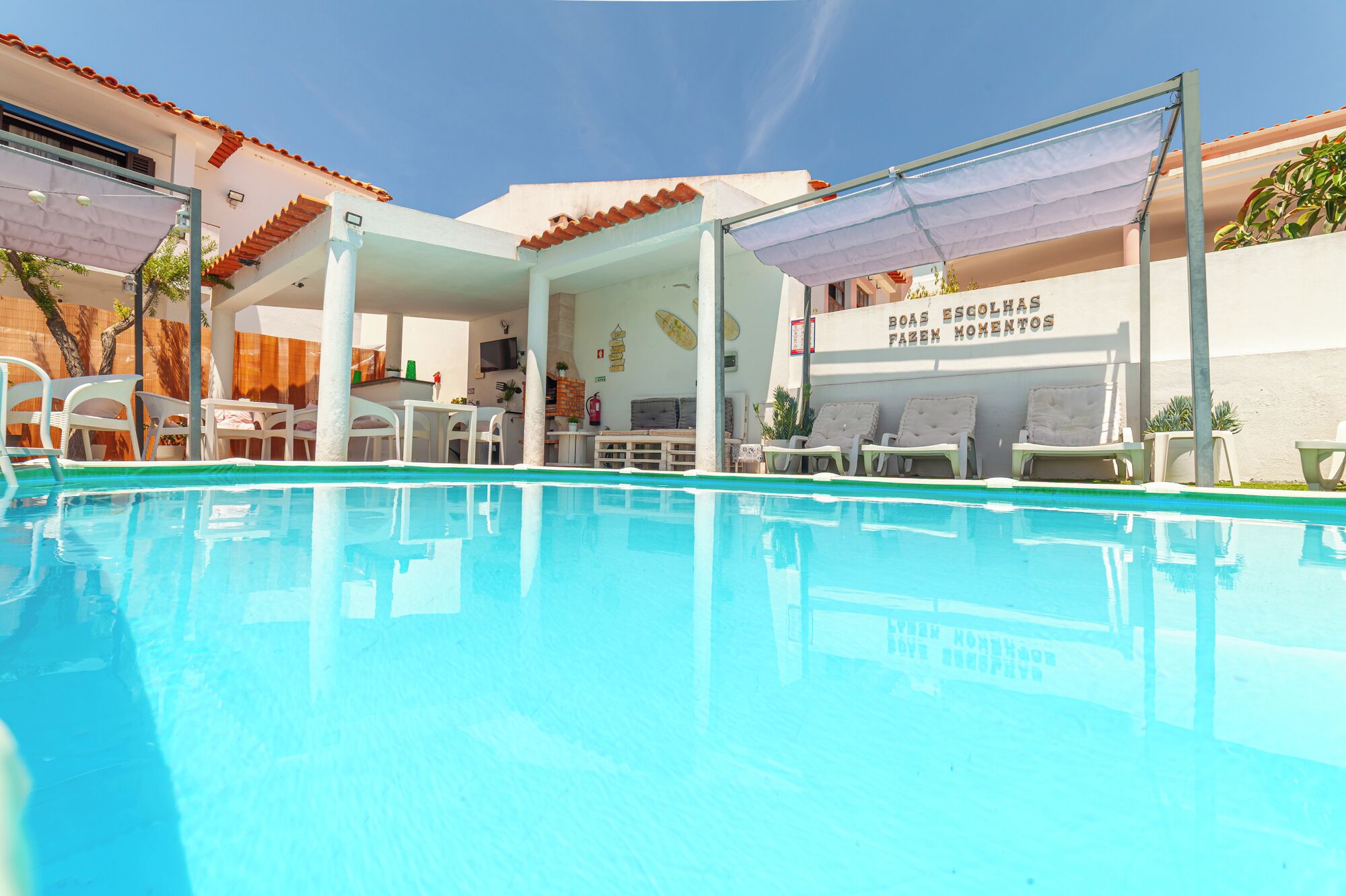 A swimming pool with lounge chairs and umbrellas, a covered seating area, and a wall with a sign.