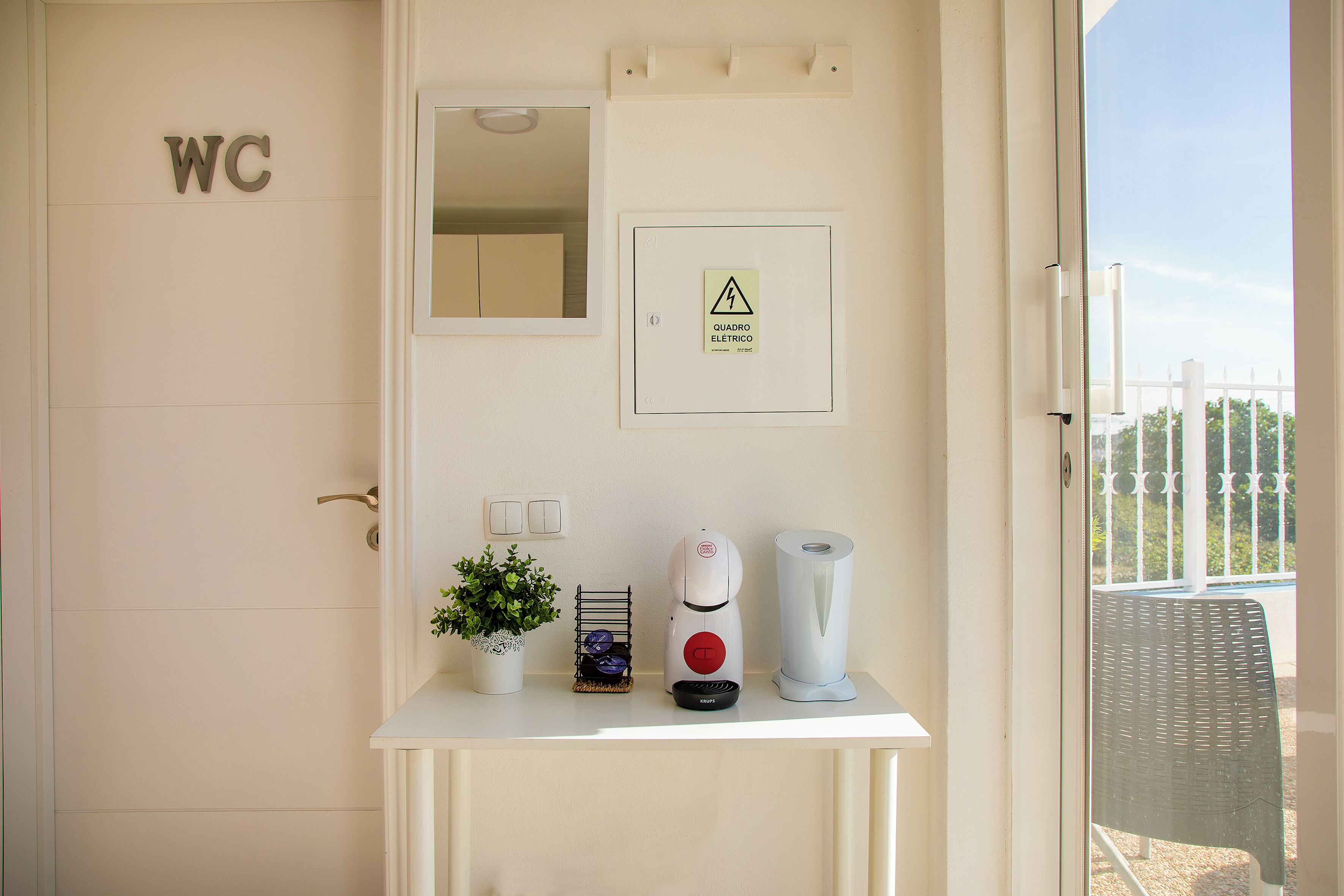 Señal de baño adaptado, señal de peligro eléctrico, una cafetera, una planta y una silla en una habitación de hotel.