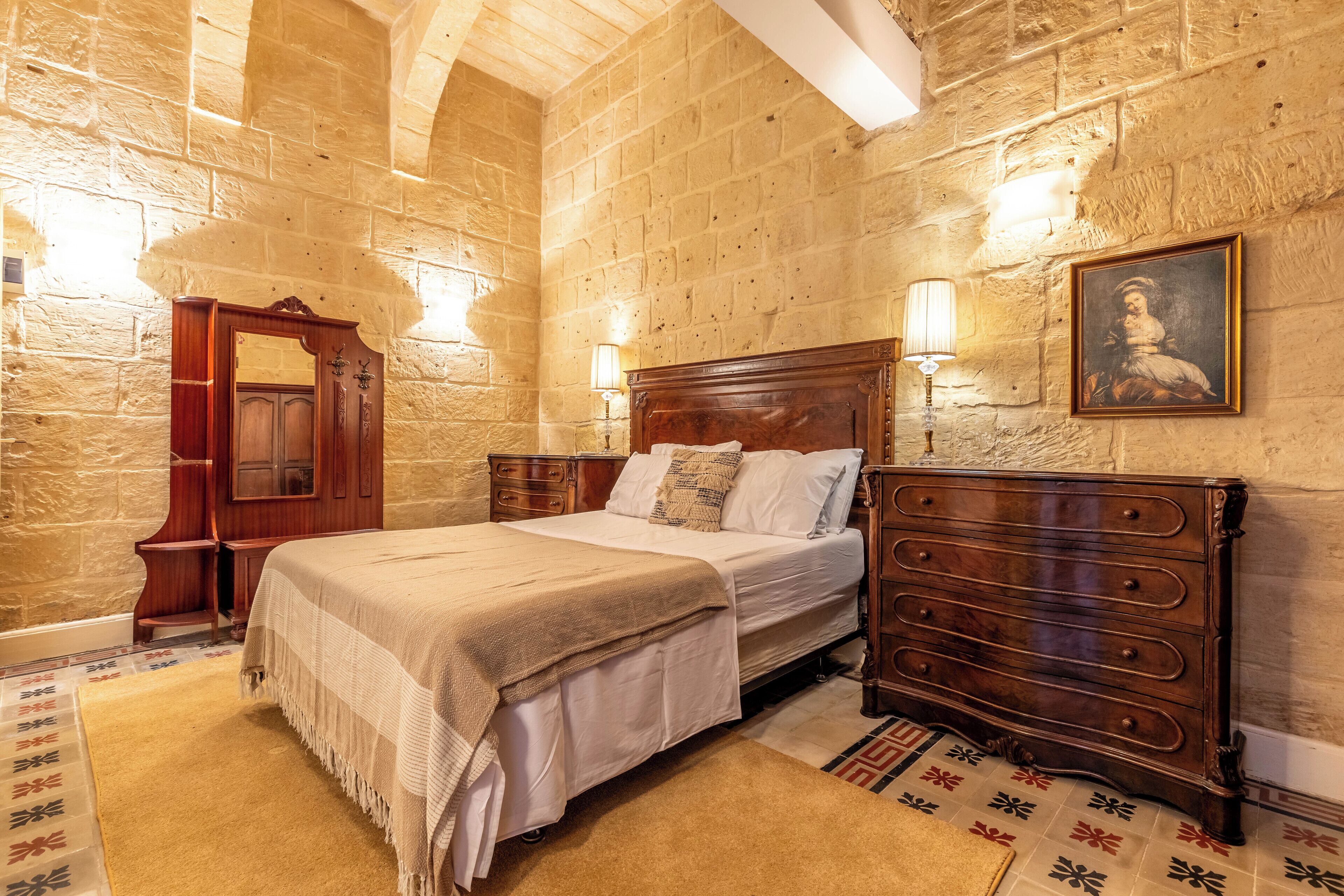 Un dormitorio con una cama grande, cómoda de madera y un espejo. La habitación tiene paredes de piedra y una alfombra estampada.