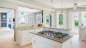 A modern kitchen with white cabinetry, a central island with a stove, and large windows offering views of greenery. - Luxurious Oceanfront Villa El Oceano with Ocean views (Grand Harbour)