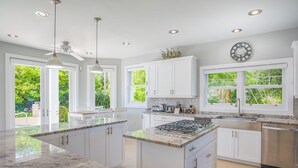 A modern kitchen with white cabinets, a central island, stainless steel appliances, and large windows with greenery outside. - Luxurious Oceanfront Villa El Oceano with Ocean views (Grand Harbour)