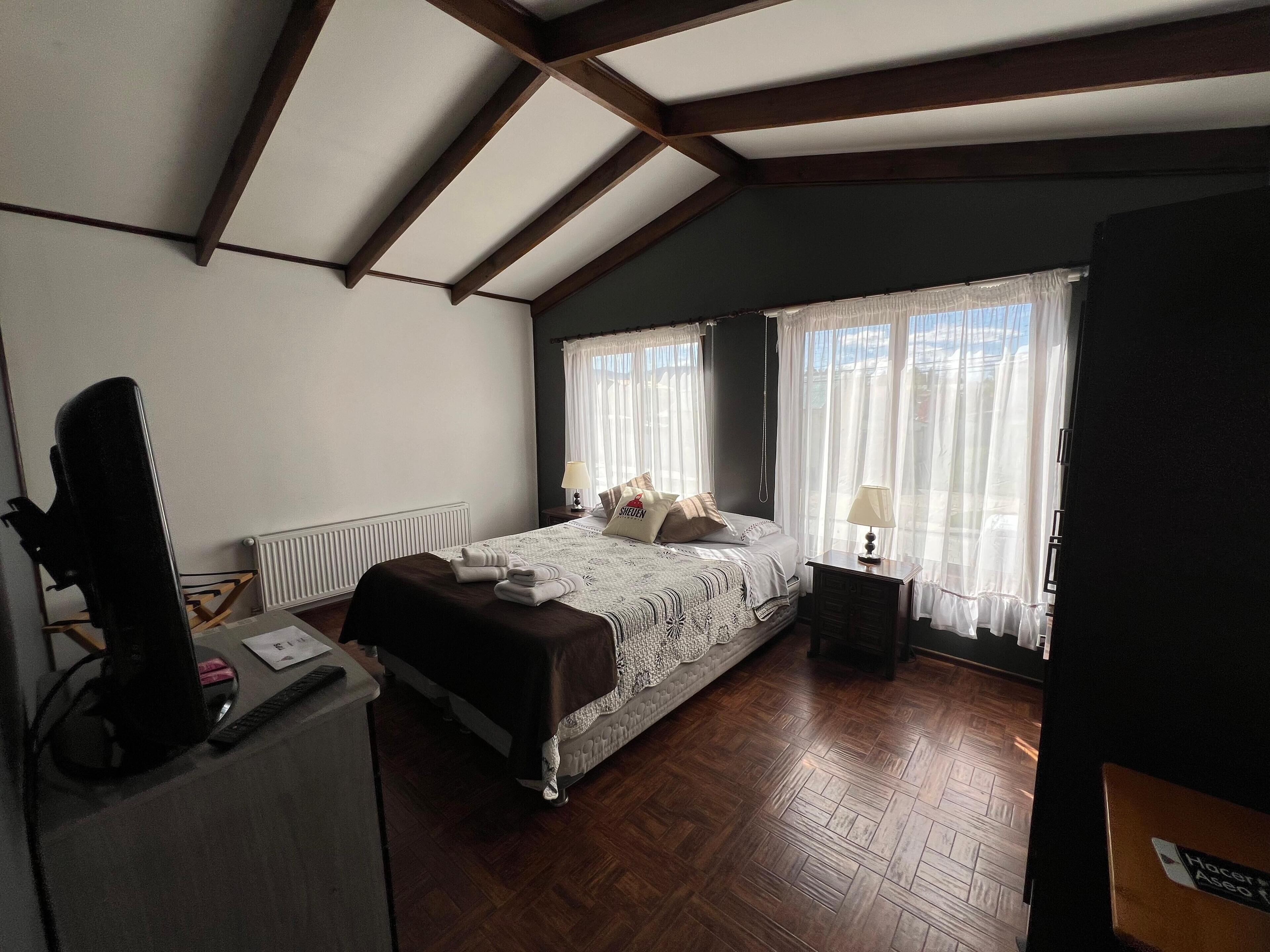Un dormitorio con suelo de madera, una cama con colcha blanca y marrón, dos lámparas de noche y una ventana con cortinas traslúcidas.
