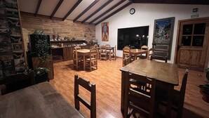 A cozy dining area with wooden tables and chairs, a kitchen in the background, and a large window. - SHEUEN PATAGONIA (Natales)