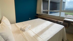 A hotel room with a bed, a desk, and a window with a view of a building and landscape. - Kariris Blue Tower (Crato)