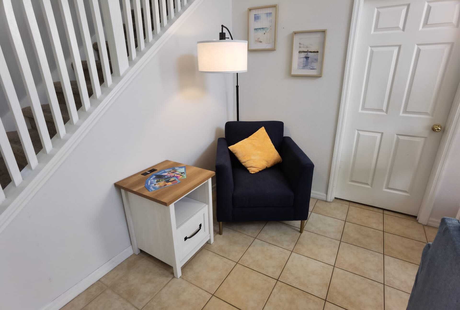 Una habitación con una escalera, una pequeña mesa con revistas, un sillón oscuro con cojín, una lámpara de pie y una puerta.