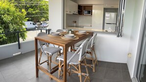 A dining area with a wooden table and wicker chairs, a view of the kitchen with modern appliances, and a balcony with a railing and greenery. - Great size close to the action (Manly)