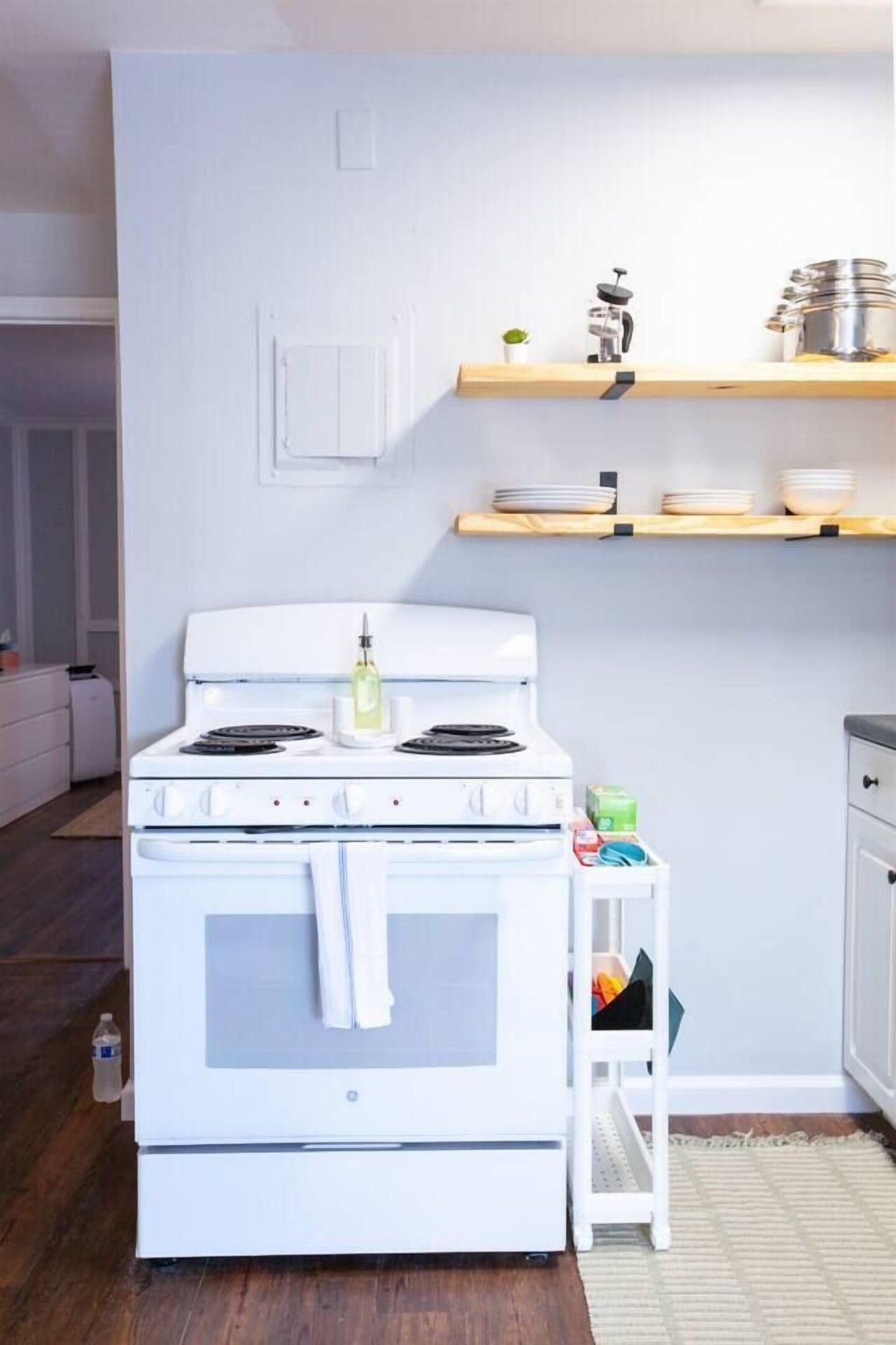 Une cuisine avec un four blanc, des étagères en bois au-dessus et une petite table sur laquelle sont posés des ustensiles de cuisine.