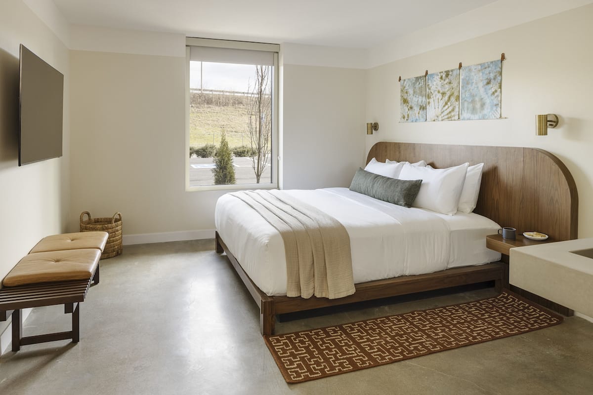 A bedroom with a large bed, a bench, a bedside table, a wall-mounted TV, and a window with a view of a field.