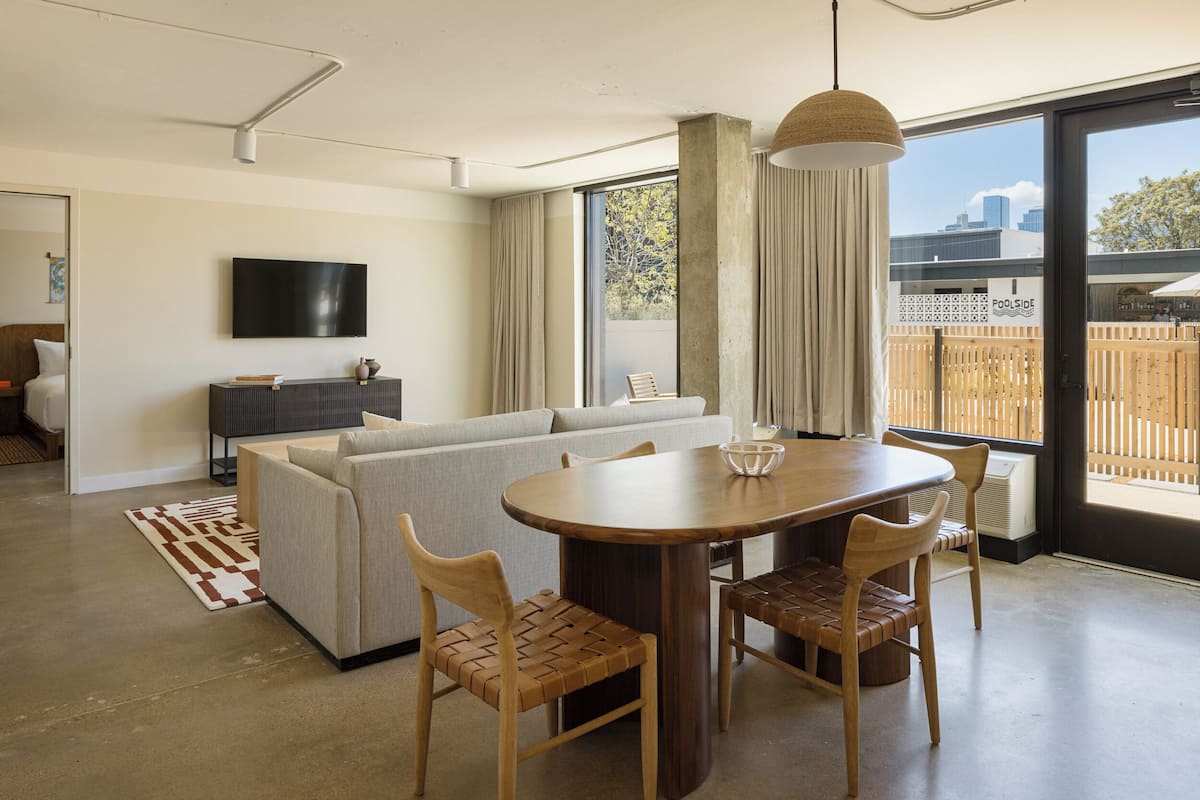 A modern living room with a sofa, dining table, and chairs. There is a TV mounted on the wall and a large window with a view of the city.