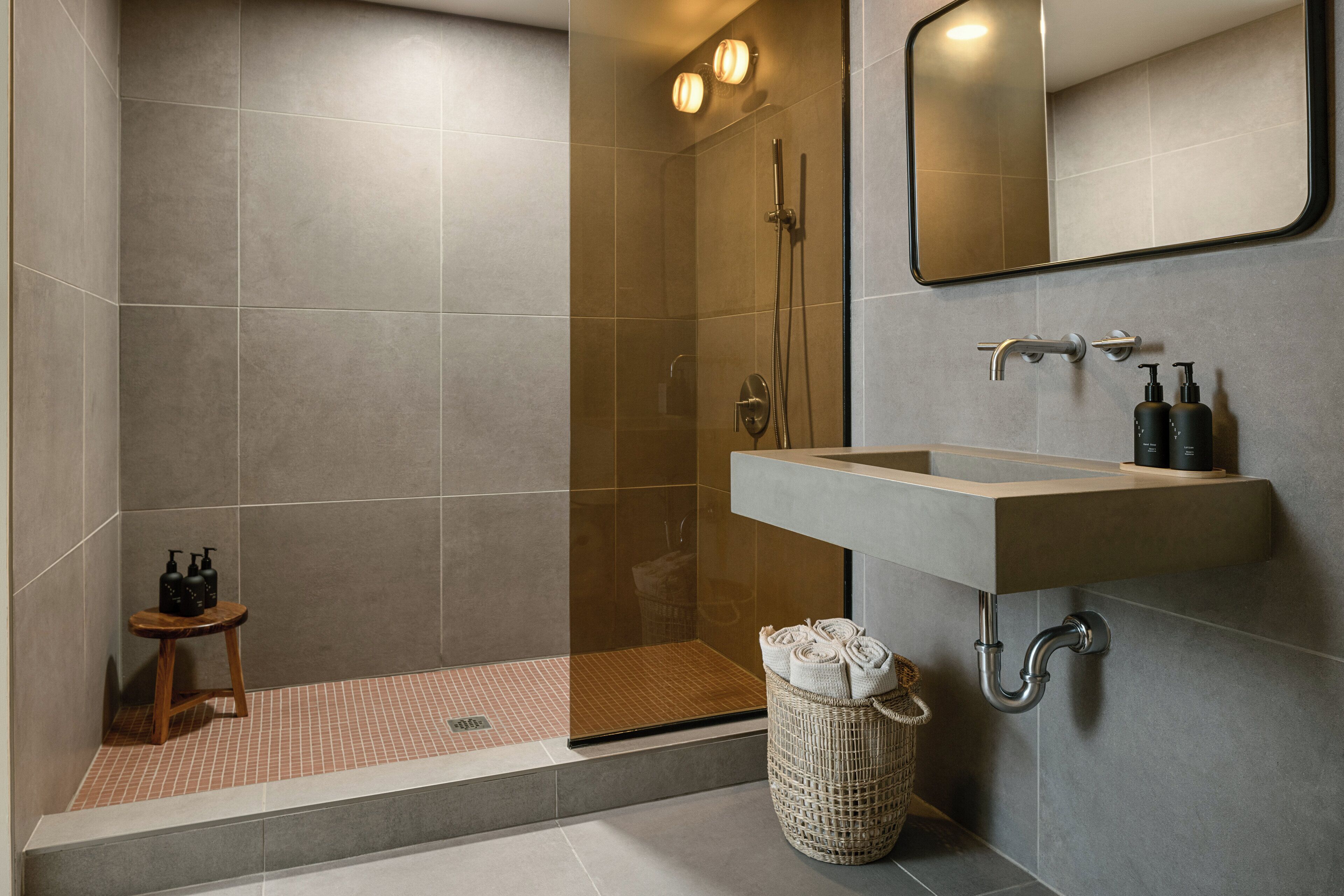 A modern bathroom with a glass-enclosed shower, a sink with a mirror above, and a small stool with toiletries.