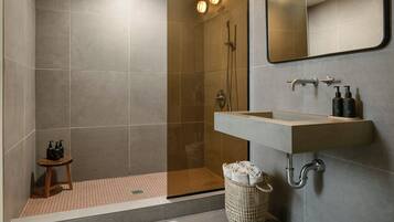 A modern bathroom with a glass-enclosed shower, a sink with a mirror above, and a small stool with toiletries.