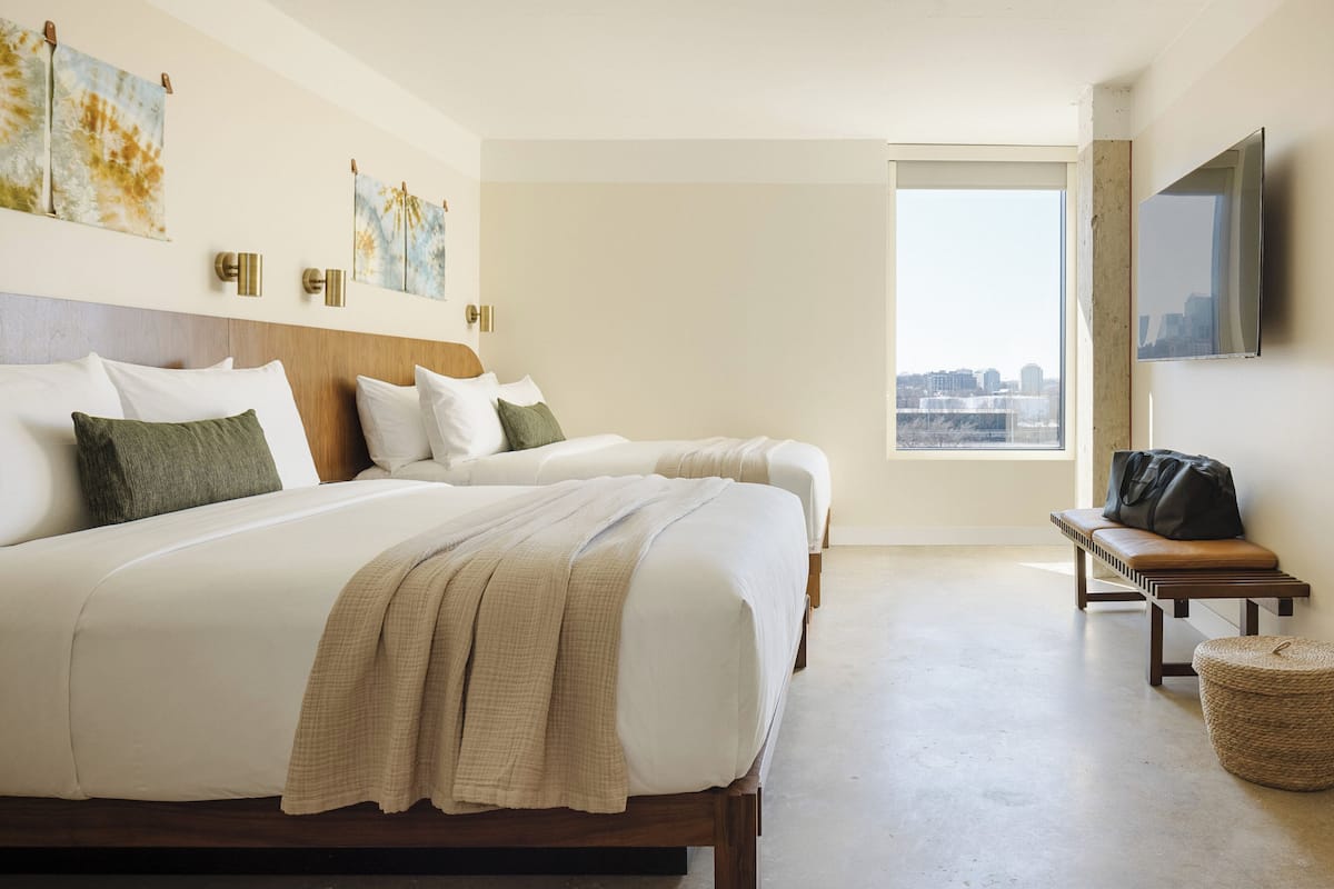 A hotel room with two beds, a large window with a city view, a flat-screen TV mounted on the wall, and a bench with a bag on it.
