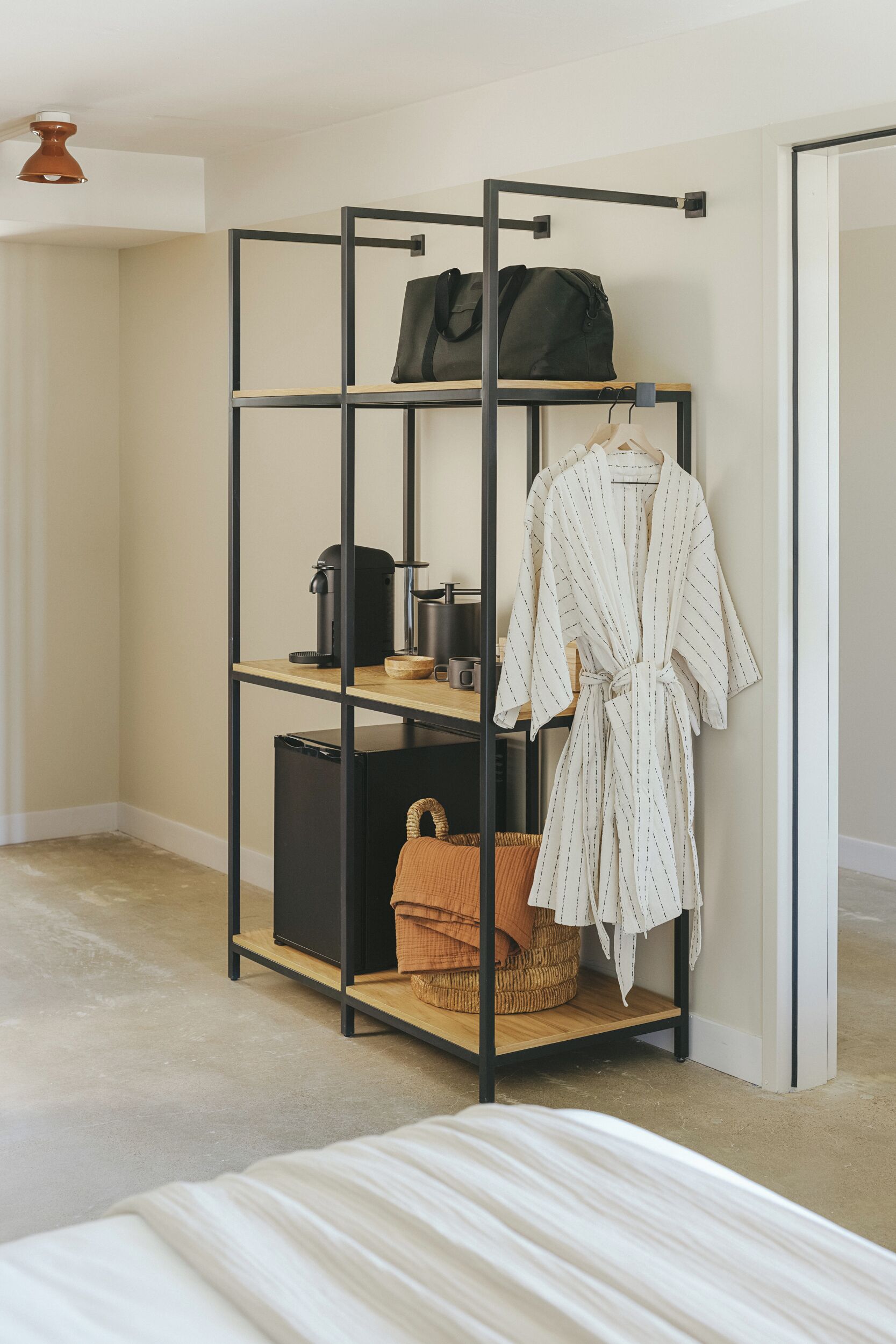 A room with a bed, a black metal shelf unit with a robe, a bag, a coffee maker, and a basket.