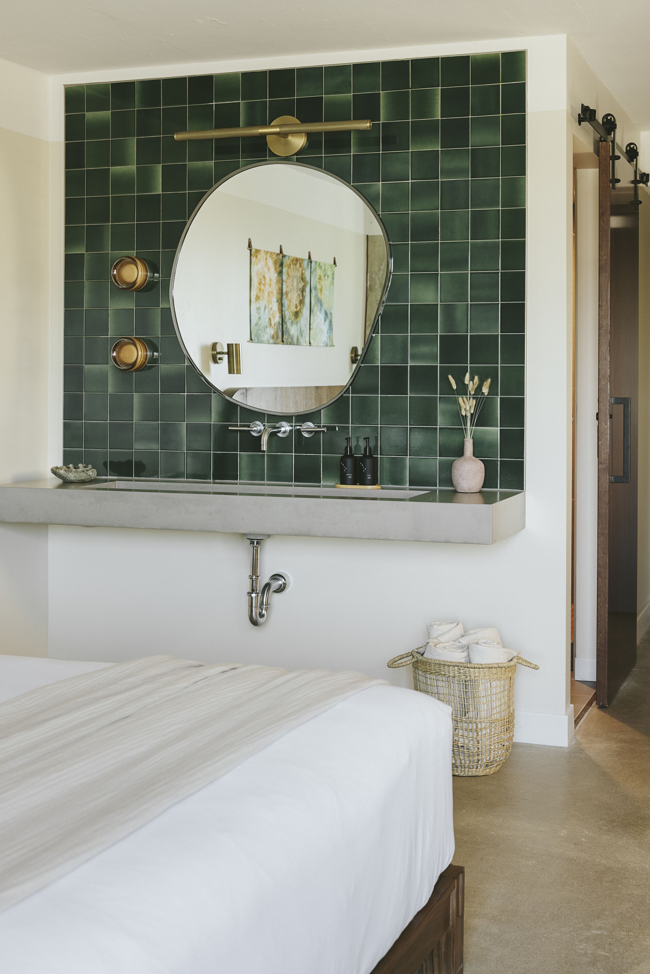 Een slaapkamer met een bed, een badkamer met een muur van groene tegels, een ronde spiegel en een wastafel.
