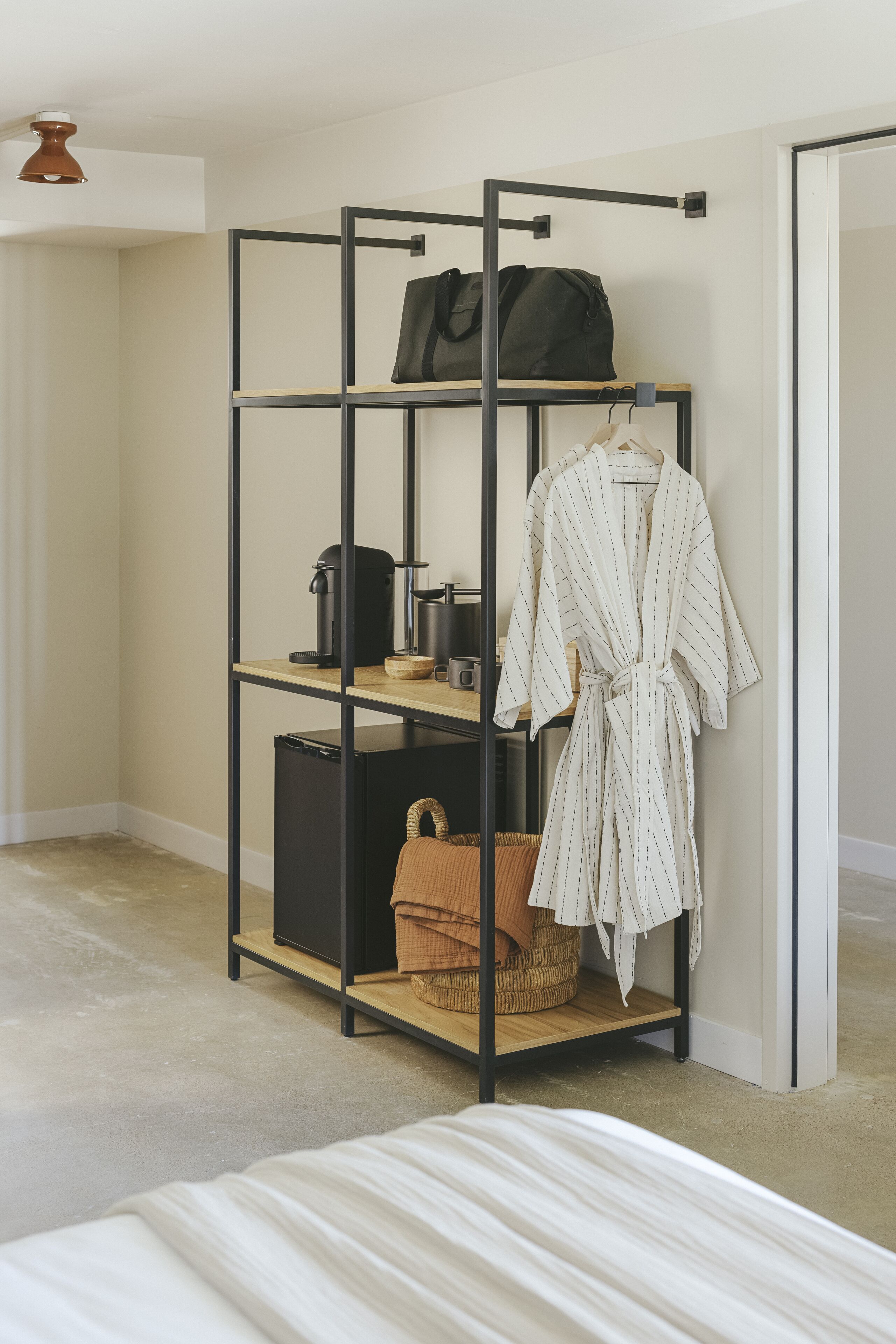 Een kamer met een bed, een zwart metalen rek met een badjas, een tas en een mand.