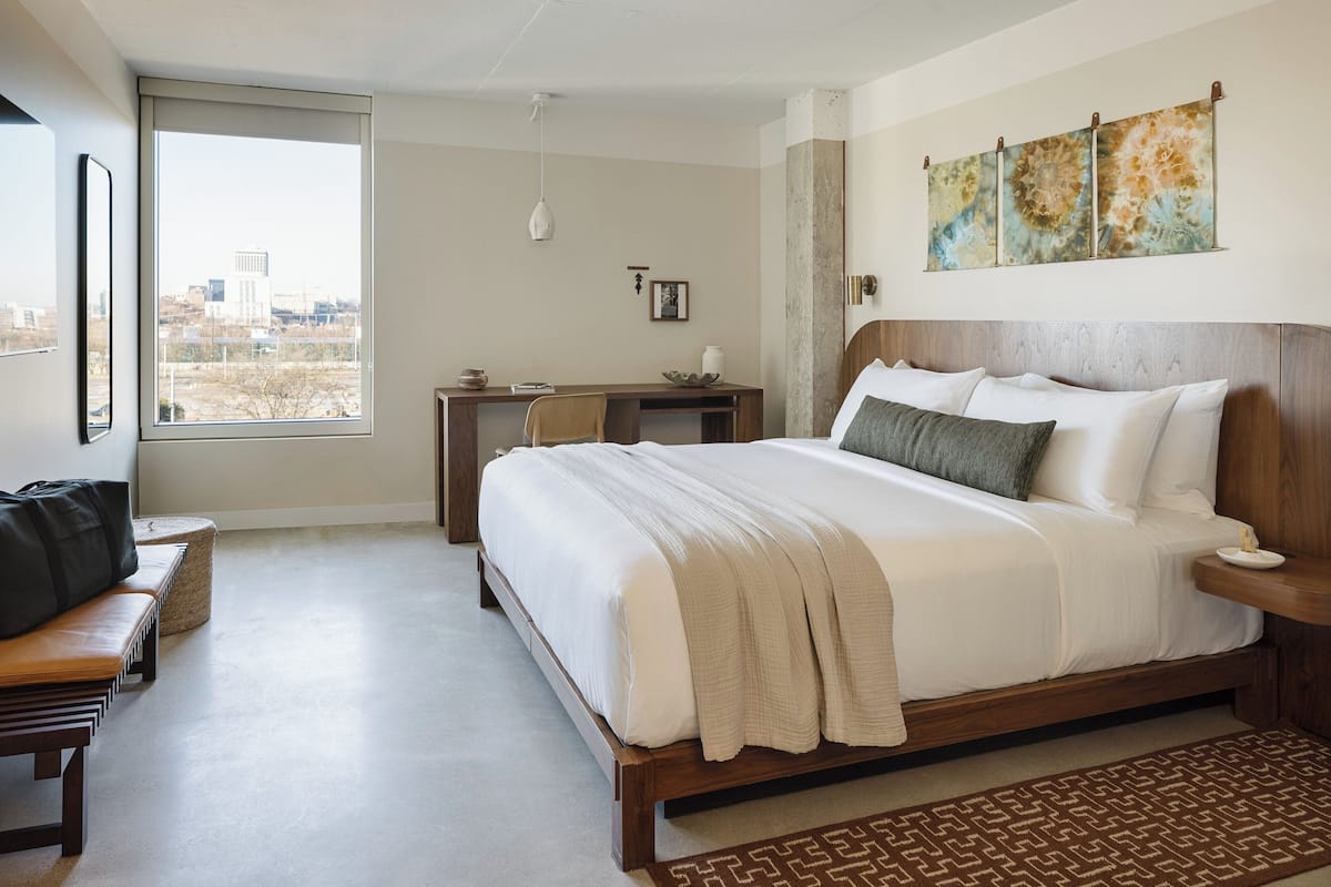 A hotel room with a large bed, a desk, a chair, a bench, and a city view.
