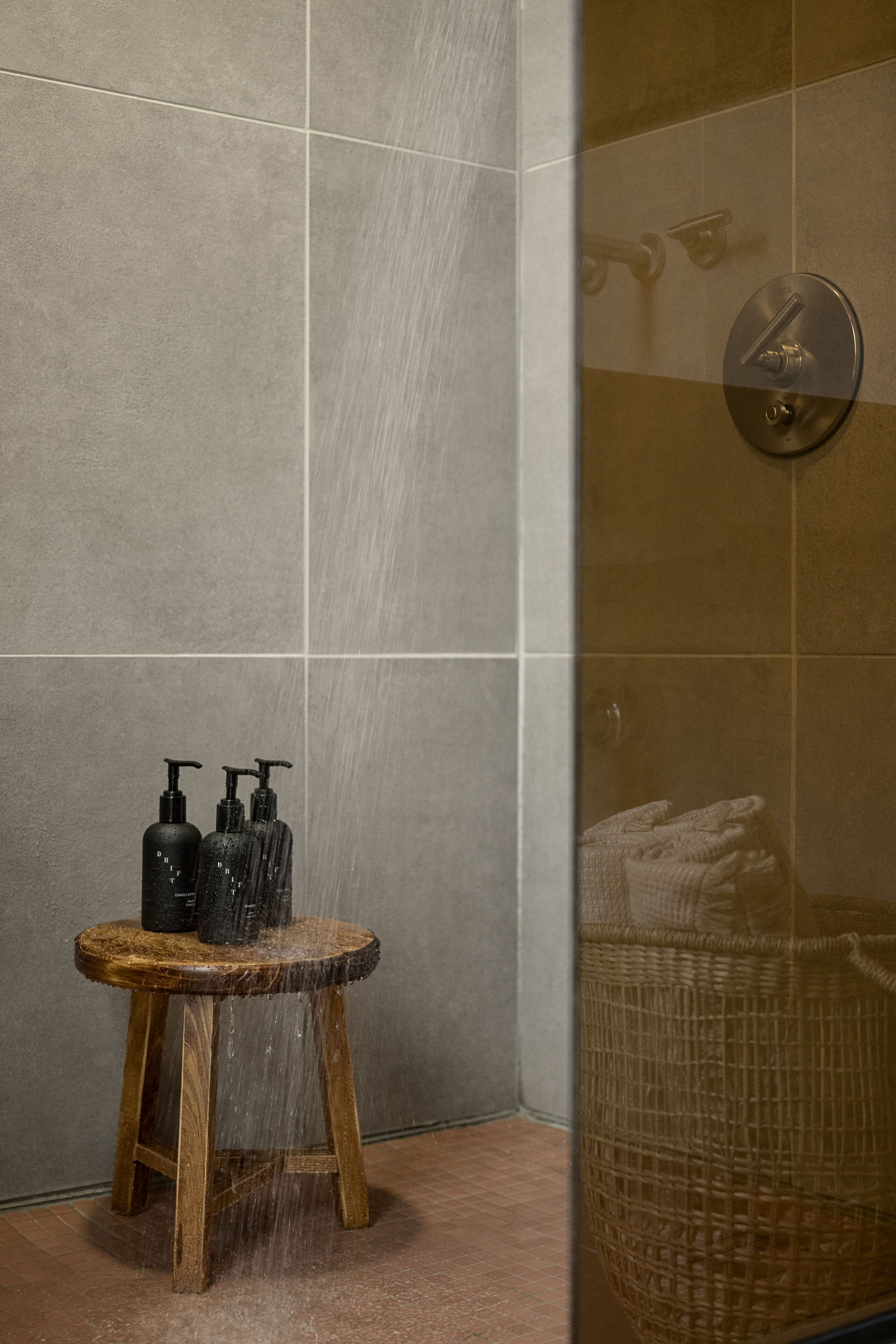 Un espace douche avec un tabouret en bois, deux bouteilles noires et un panier tressé.