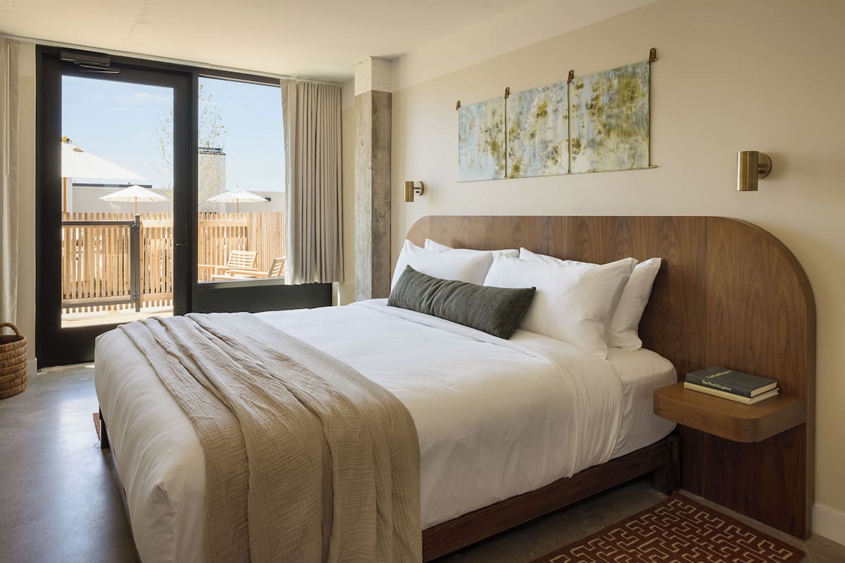 A bedroom with a large bed, a wooden headboard, a bedside table with a book, and a view of the outdoors through a sliding door.