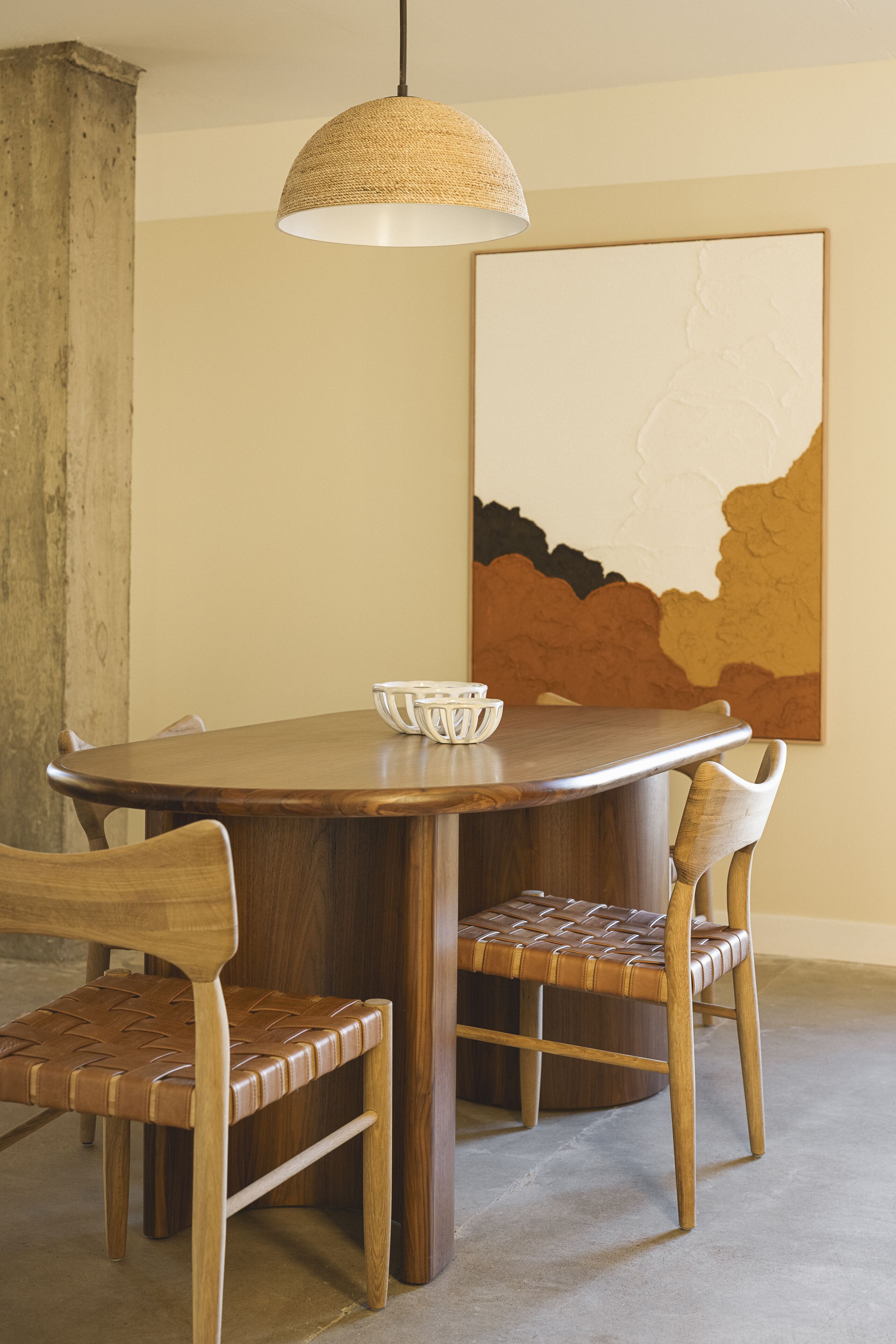 Un comedor con una mesa de madera, dos sillas, una lámpara colgante de fibras tejidas y un cuadro abstracto grande en la pared.