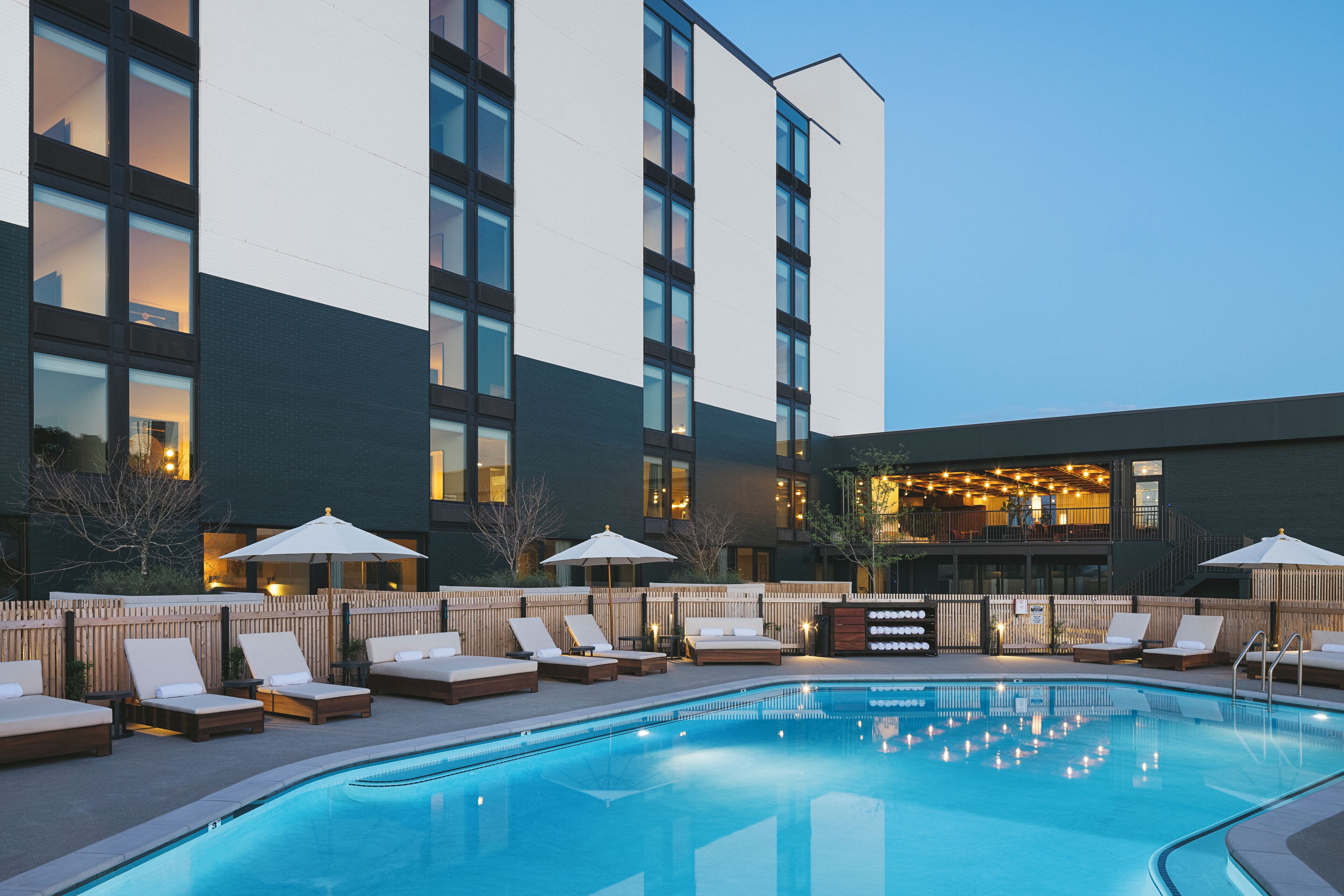 Zona de piscina de hotel con sillones de descanso y sombrillas, rodeada por una cerca de madera y un edificio con varias ventanas.