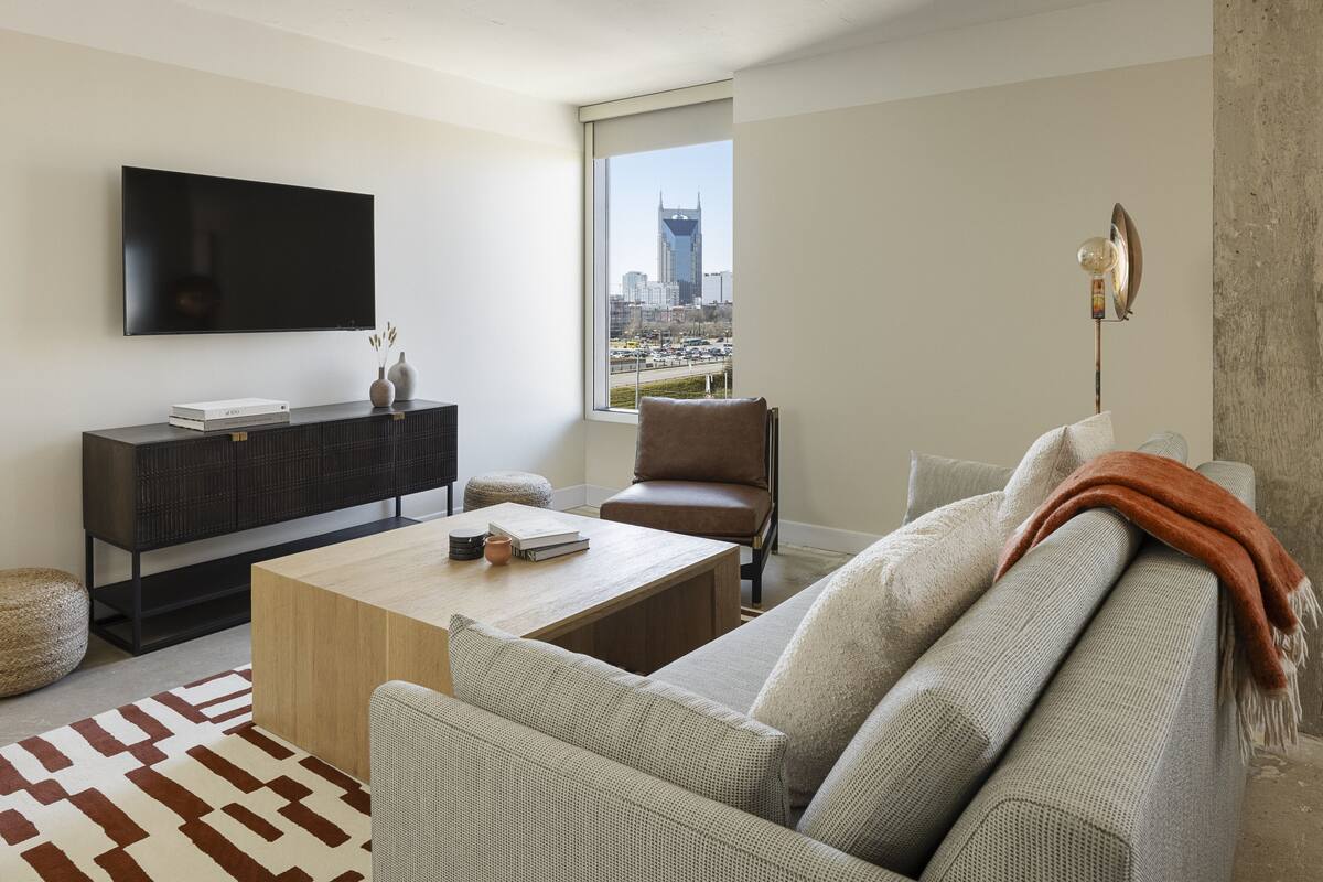 A modern living room with a flat-screen TV, a wooden coffee table, a grey sofa, and a view of a cityscape through a large window.