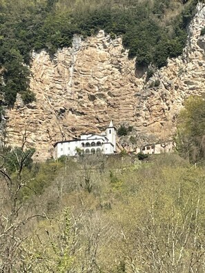 Un bâtiment blanc surmonté d’un clocher, entouré de falaises rocheuses et d’une végétation clairsemée.