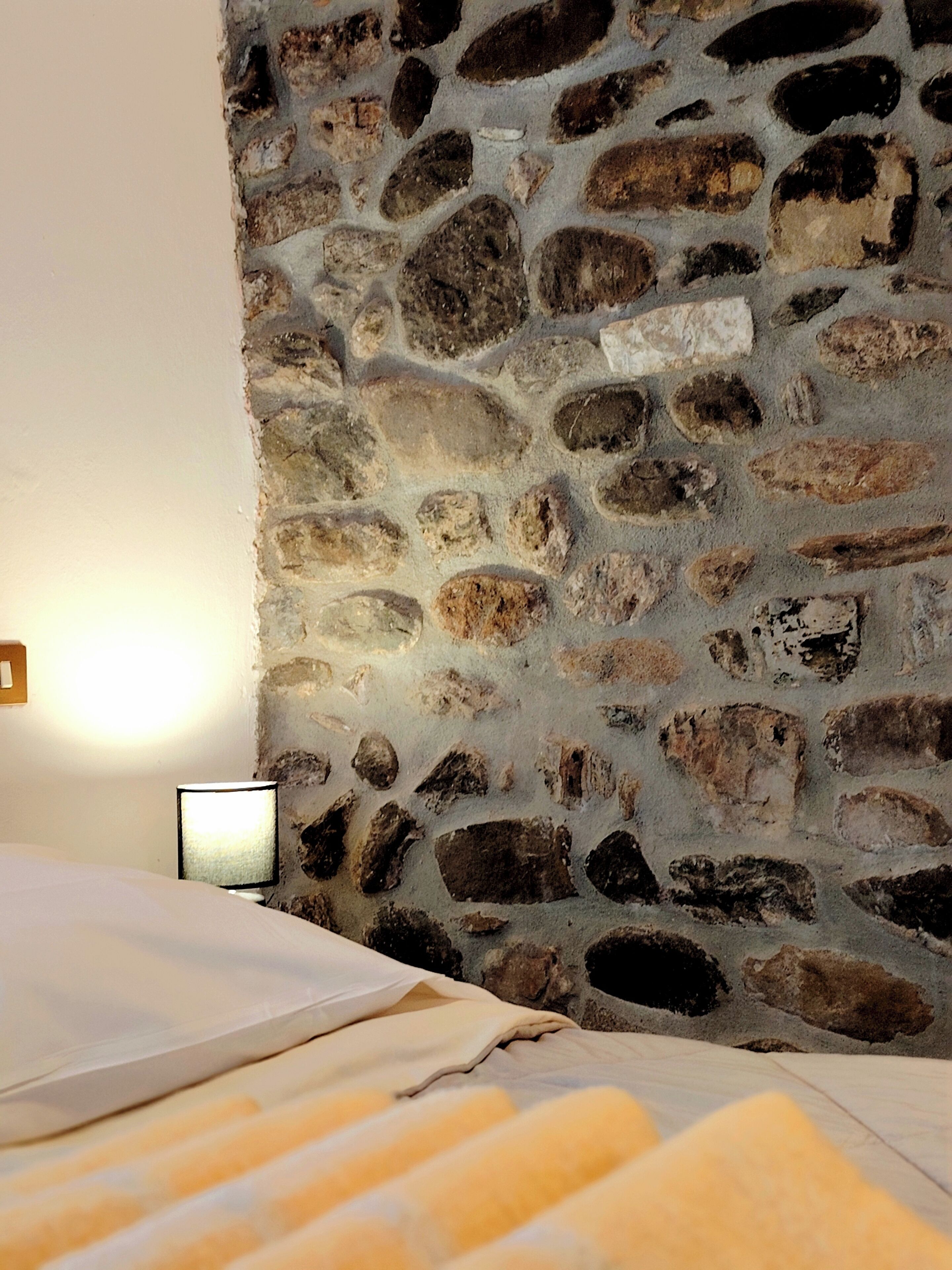 Een kamer met een stenen muur, een bed met een witte dekbedovertrek en een bedlampje.