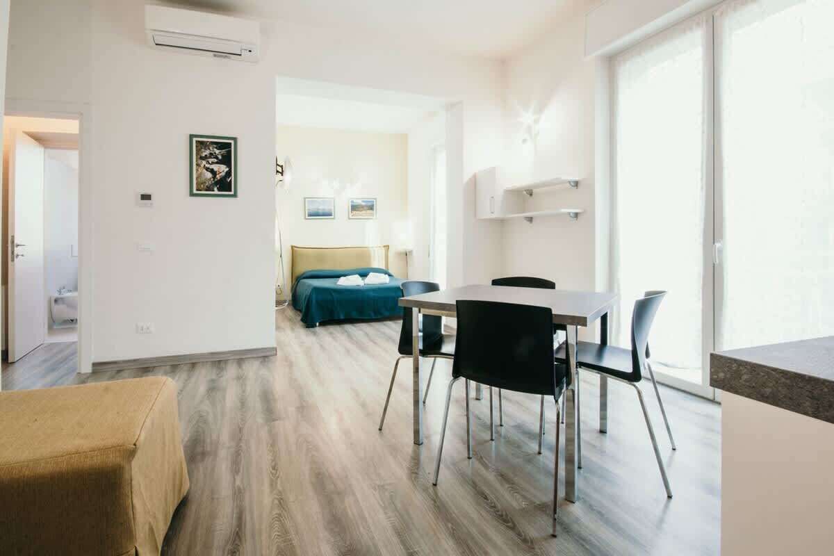 Une chambre d’hôtel moderne avec un plancher en bois, un lit recouvert d’une courtepointe bleue, un coin repas avec une table et des chaises, et une grande fenêtre laissant entrer la lumière naturelle.