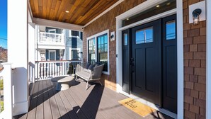 Una terraza de madera con barandal blanco, puerta negra y banco con cojines.