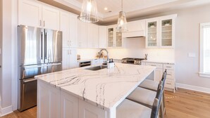 Una cocina moderna con armarios blancos, una isla grande de mármol, electrodomésticos de acero inoxidable e iluminación colgante.