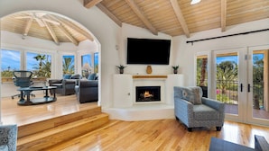 Un amplio salón con chimenea, asientos cómodos y ventanas grandes que ofrecen vistas a palmeras.