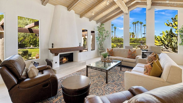 Un salón espacioso con chimenea, sofás de cuero, una mesa de centro y ventanas amplias que dan a un jardín y palmeras.