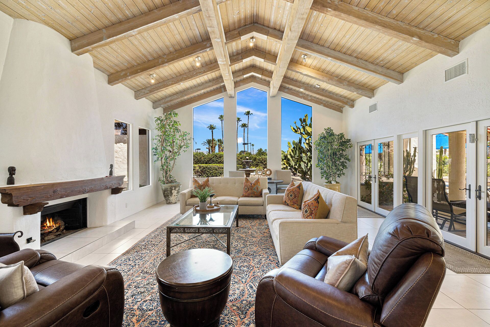 A spacious living room with a high wooden ceiling, a fireplace, a large sofa set, and a view of palm trees through large windows.