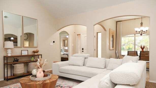 Ein Wohnzimmer mit einem weißen Sofa, einem Holztisch und einem farbenfrohen Teppich. An den Wänden befinden sich zwei Spiegel, eine Chandelier und ein Fenster mit Jalousien.