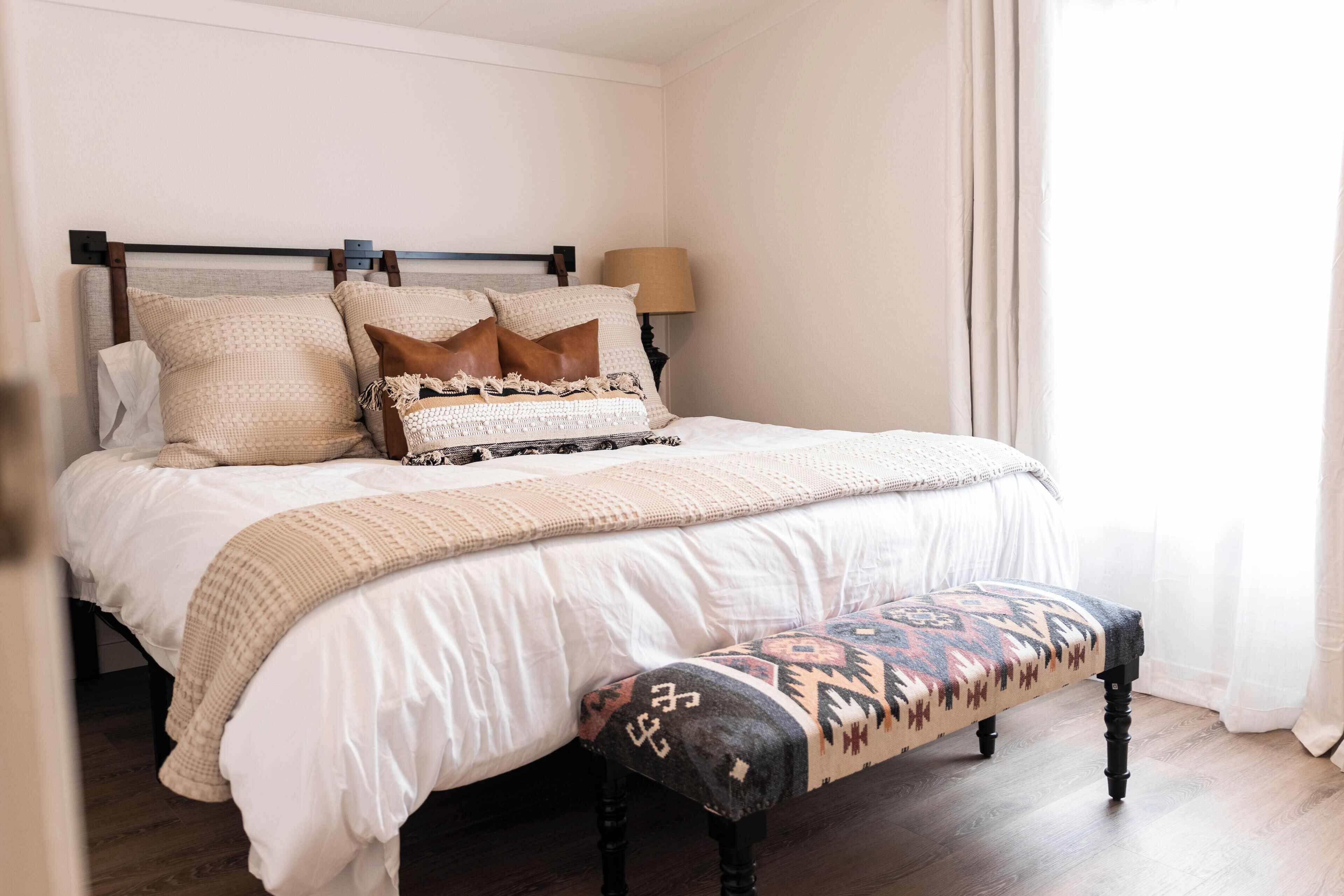 Una cama bien hecha con una colcha y almohadas estampadas, una lámpara de noche y un banco con un diseño geométrico.