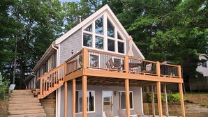 Ein zweistöckiges Haus mit einer Holzdachterrasse und einer Sitzgelegenheit im Freien.